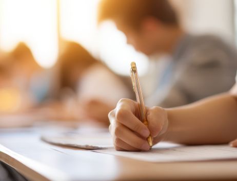 Nahaufnahme einer Person, die mit einem Stift eine Prüfung oder Klassenarbeit schreibt, im unscharfen Hintergrund weitere sitzende Teilnehmende in einem Unterrichtsraum