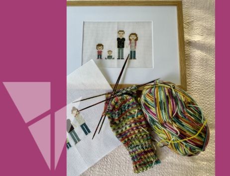 Ein Stickbild einer Familie in einem Rahmen, daneben ein gestrickter Socken und ein Knäuel bunter Wolle.