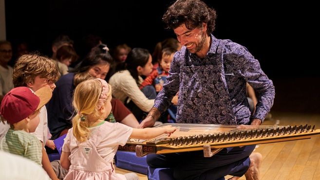 Ein Musiker spielt ein Saiteninstrument vor einer Gruppe von Kindern, die fasziniert zuschauen und zuhören.