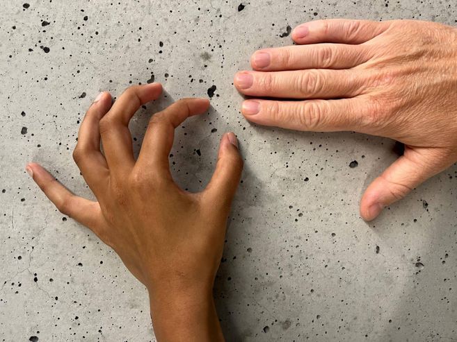 Das BIld zeigt zwei Hände. Oben im Rechten Bildrandliegt eine glatte Hand auf einer Oberfläche aus Beton. Unten mittig ragt eine linke Hand ins Bild hinein. Die Finger formen jeweils angedeutete Kreise.
