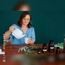 Eine Frau gießt Tee aus einer weißen Kanne in eine Tasse. Auf dem Tisch stehen Kräuter und Fläschchen.