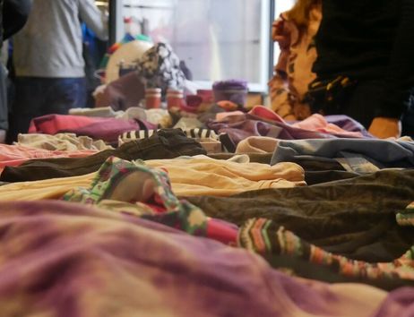 Tisch mit ausgebreiteten Kleidungsstücken auf einem Flohmarkt, im Hintergrund stehen mehrere Personen.