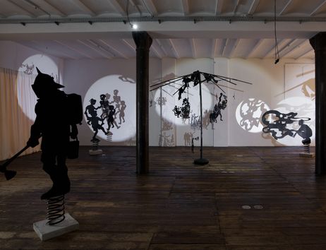 Installationsansicht der Ausstellung Keiner rannte von Andrey Klassen. In einem Raum mit zwei Säulen steht im Vordergrund eine große Figur, der Feuerbändiger. Im Hintergrund sind verbrannte Sonnenschirme, weitere Figuren und Projektionen zu sehen.