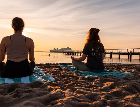 Zwei Personen meditieren am Strand bei Sonnenuntergang mit einem Schiff im Hintergrund.