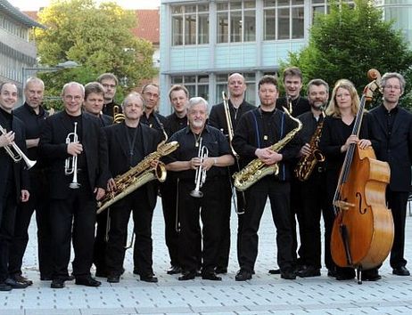 Eine Jazzband posiert mit ihren Instrumenten im Freien vor einem modernen Gebäude.