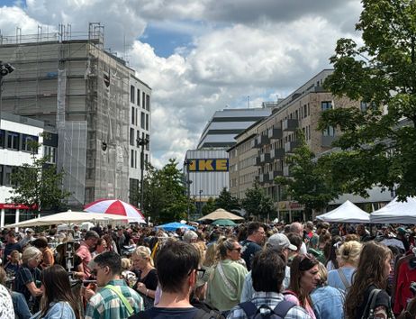 Viele Menschen im unteren Teil des Bildes, befinden sich auf einem Flohmarkt draußen in der Sonne. Im Hintergrund sind Gebäude zu sehen und ganz am hinten im Bilde der IKEA in der Neuen Grossen Bergstrasse in Altona.