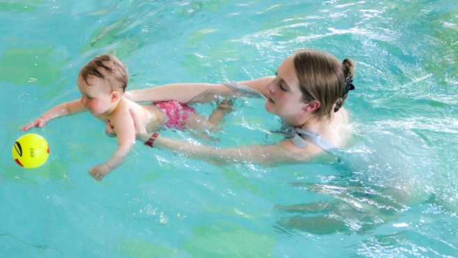 Eine Frau hält ein Baby im Wasser, während es nach einem gelben Ball greift.