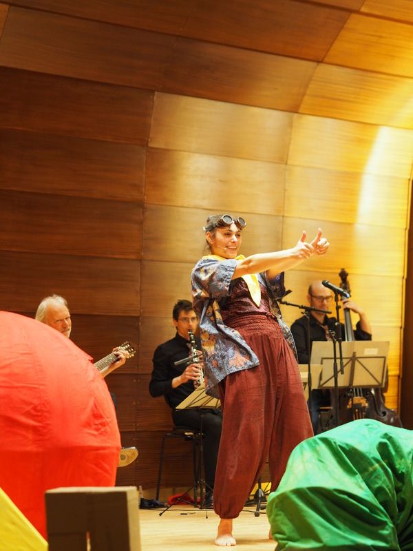 Eine Person in bunter Kleidung führt auf einer Bühne vor einem kleinen Orchester eine Theateraufführung auf.