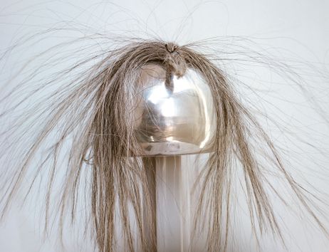 Das Bild zeigt einen Van-de-Graaff-Generator, vor weißem Hintergrund, auf dem sehr dicke graue Haare zusammengebunden liegen, ein paar einzelne Haare stehen hoch.