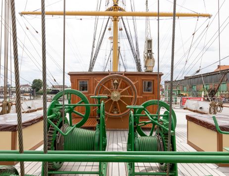 Deck eines historischen Segelschiffs mit Steuerrad und grüner Winde.