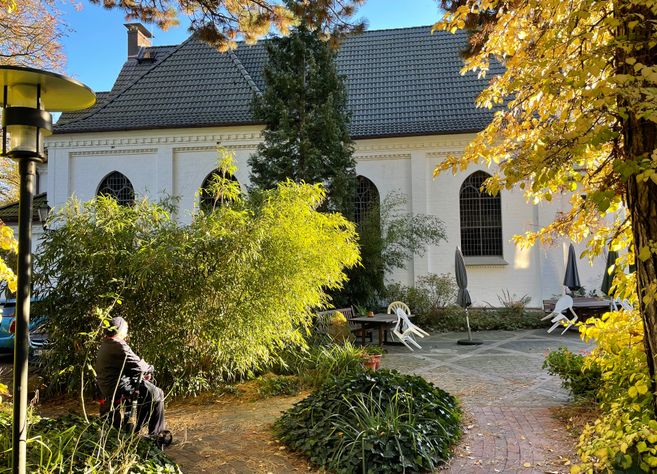 Ein weißes Gebäude mit großen Fenstern, umgeben von herbstlichen Bäumen und einem kleinen Garten mit Sitzgelegenheiten.