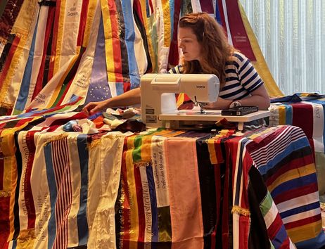Eine Frau näht an einer Nähmaschine an einem großen, bunten Vorhang aus vielen bedruckten Stoffbändern mit Erinnerungs- und Gedenktexten.
