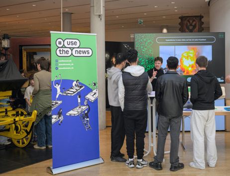 Gruppe von Menschen bei einer Präsentation mit einem großen Bildschirm und einem Banner mit der Aufschrift '#use the news'.