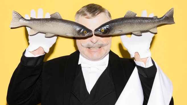Ein Mann in einem weißen Hemd, weißen Handschuhen und einem schwarzen Jacket steht vor einem gelben Hintergrund. Er hält in jeder Hand einen Fisch, dessen Köpfe vor seinen eigenen Augen liegen.