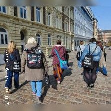 Gruppe von Menschen mit Einkaufstaschen geht auf einer gepflasterten Straße entlang historischer Gebäude.