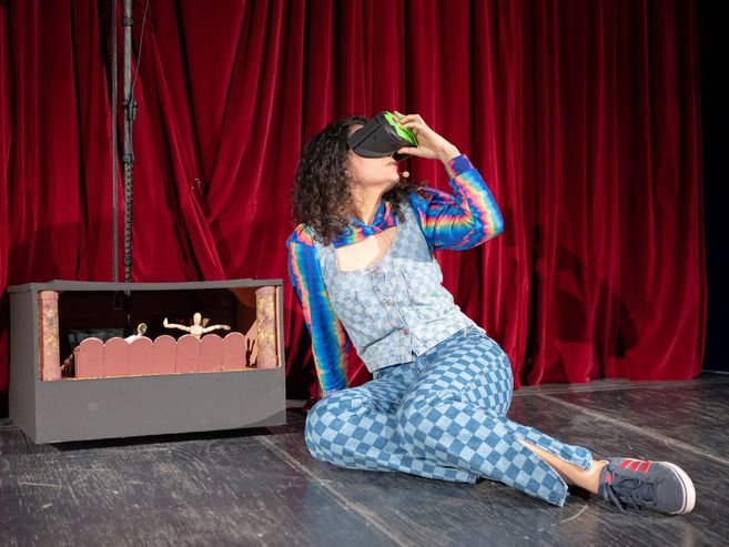 A woman dressed in colourful clothes is sitting on a stage in front of a red velvet curtain, holding a pair of VR glasses up to her eyes.