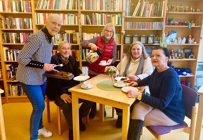 Eine Gruppe älterer Menschen sitzt an einem Tisch in einem Café mit Bücherregalen im Hintergrund. Eine Frau serviert Kuchen und Kaffee.