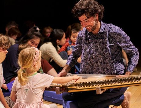 Ein Musiker spielt ein Saiteninstrument vor einer Gruppe von Kindern, die fasziniert zuschauen und zuhören.