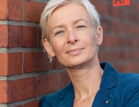Porträt einer Frau mit kurzem blondem Haar, die an eine Backsteinmauer lehnt. Sie trägt einen blauen Blazer. Oben rechts ist ein rotes Logo mit den Buchstaben 'HF'.