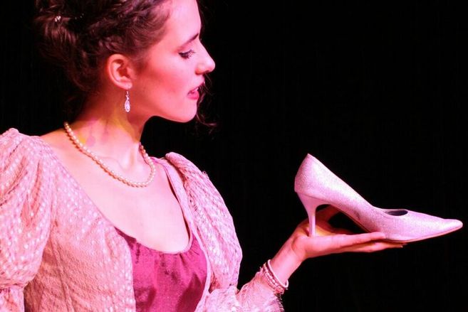 Eine junge Frau im Kostüm hält einen glitzernden rosa High Heel in der Hand, passend zum Märchen-Musical Aschenputtel.