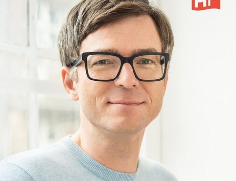 Porträt eines Mannes mit Brille und hellblauem Pullover vor einem Fenster.