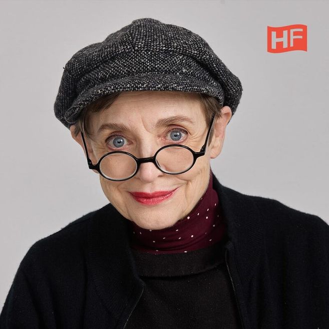 Eine ältere Frau mit Brille und Mütze lächelt in die Kamera. Sie trägt roten Lippenstift und einen schwarzen Pullover.