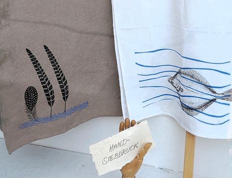 Two hand-printed dish towels hang side by side. The left towel displays stylized plants, while the right towel depicts a fish in blue waves.