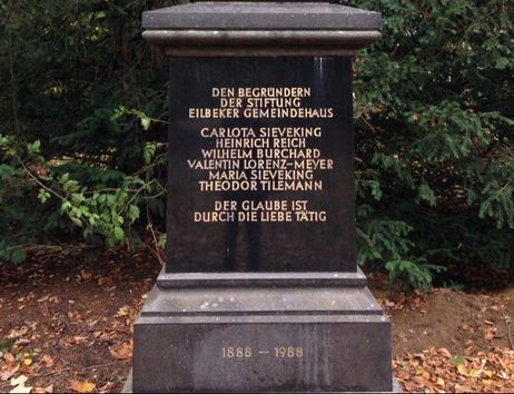 Ein schwarzer Gedenkstein mit goldener Inschrift im Jacobipark, umgeben von Bäumen und Laub.