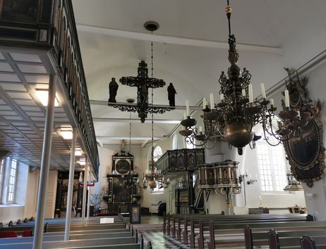 Innenansicht einer Kirche mit Emporen, Kronleuchtern, Kreuz und Altarraum; leere Bänke im Vordergrund.
