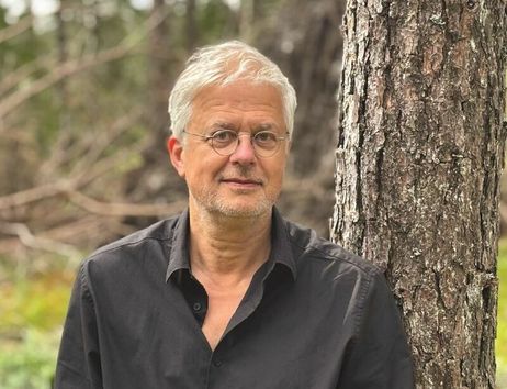 Porträt eines älteren Mannes mit grauen Haaren und Brille, der im Wald neben einem Baum steht.