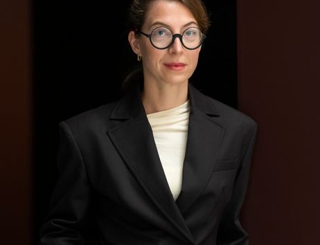 Eine Frau mit Brille und schwarzem Blazer steht vor einem dunklen Hintergrund.