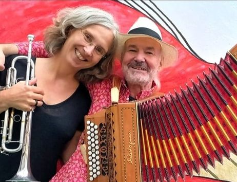 Ein Mann mit Akkordeon und eine Frau mit Trompete lächeln in die Kamera vor einem bunten Hintergrund.