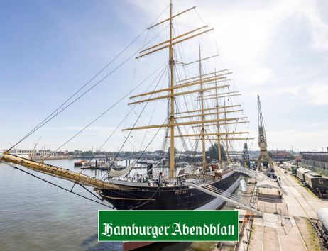 Das Segelschiff Peking liegt im Hafen, umgeben von Kränen und Gebäuden, unter einem klaren blauen Himmel.