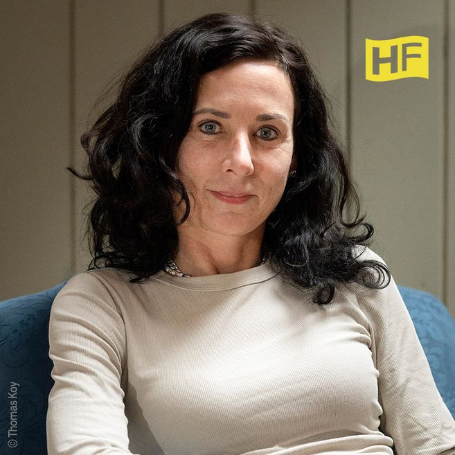Porträt einer Frau mit dunklen Haaren in einem beigen Oberteil, sitzend auf einem blauen Stuhl. Im Hintergrund ist ein HF-Logo zu sehen.