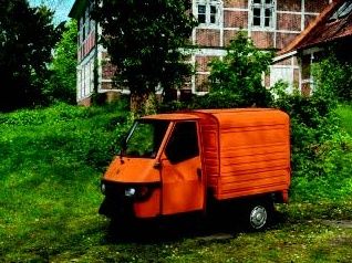 Ein kleines, orangefarbenes Dreiradfahrzeug steht auf einer grünen Wiese vor einem alten Backsteingebäude.