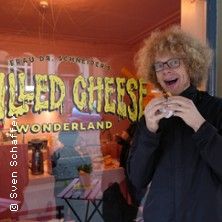 Person mit lockigem Haar isst vor einem Laden mit der Aufschrift 'Grilled Cheese Wonderland'.