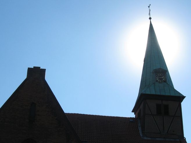 Kirchturm mit Uhr vor strahlend blauem Himmel und Sonne im Hintergrund.