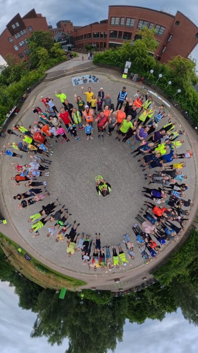 Gruppe von Menschen steht im Kreis auf einem Platz, umgeben von Gebäuden und Bäumen.