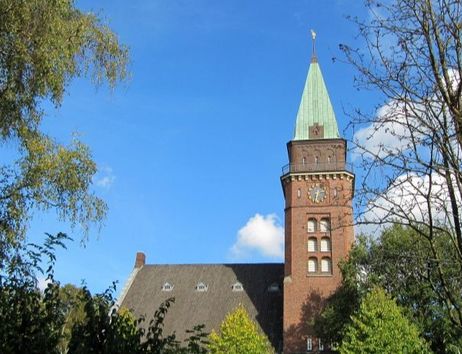 Ein Kirchturm mit grünem Dach und Uhr, umgeben von Bäumen, unter einem blauen Himmel.