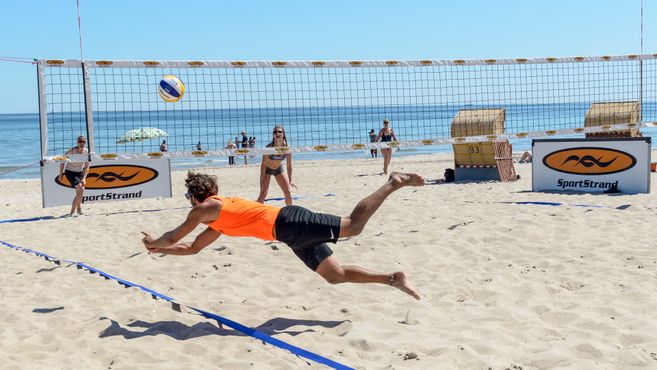Ein Mann in einem orangefarbenen Shirt springt nach einem Volleyball am Strand. Im Hintergrund sind ein Netz, Strandkörbe und das Meer zu sehen.