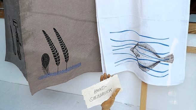 Two hand-printed dish towels hang side by side. The left towel displays stylized plants, while the right towel depicts a fish in blue waves.