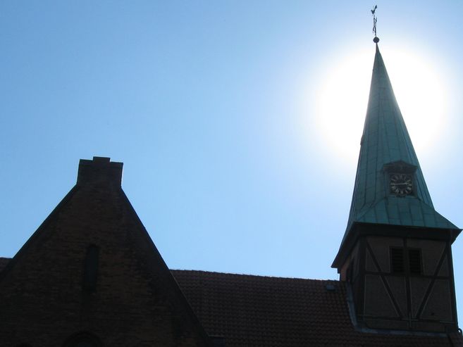Kirchturm mit Uhr vor strahlend blauem Himmel und Sonne im Hintergrund.