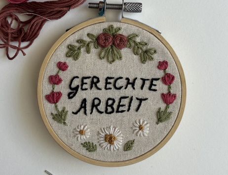 Ein Stickrahmen mit der Aufschrift 'Gerechte Arbeit', umgeben von gestickten Blumen und Blättern.