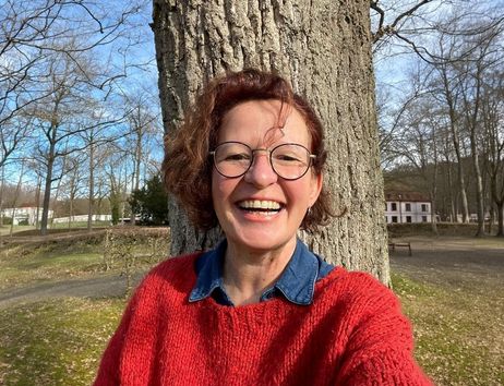 Eine lächelnde Person mit Brille und rotem Pullover steht vor einem Baum in einem Park.