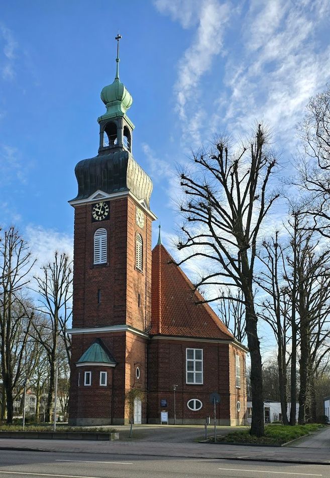 Backsteinkirche mit grünem Turm und Uhr, umgeben von kahlen Bäumen, unter blauem Himmel.