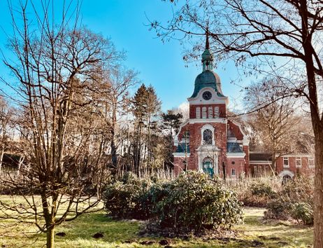 Eine rote Backsteinkirche mit grünem Turm, umgeben von Bäumen und Sträuchern im Frühling.