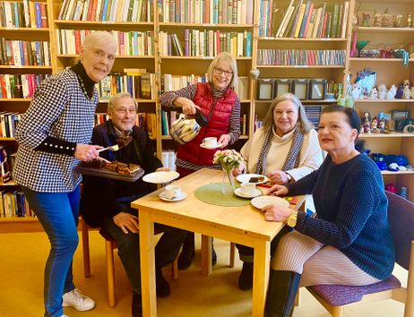 Eine Gruppe älterer Menschen sitzt in einem Café mit Bücherregalen im Hintergrund. Eine Frau serviert Kuchen und Kaffee.