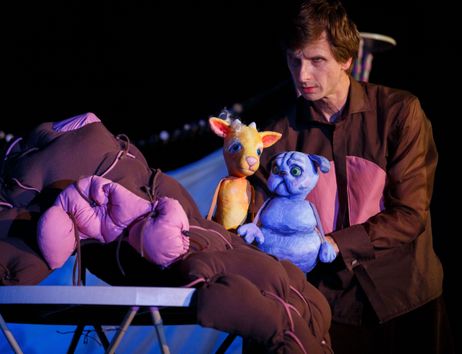 Ein Puppenspieler hält zwei bunte Puppen, eine gelbe und eine blaue, vor einem dunklen Hintergrund.