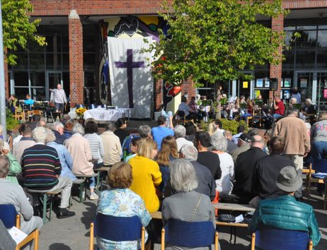 Menschen versammeln sich zu einem Open-Air-Gottesdienst vor einem Gebäude mit einem großen Kreuz auf einem Tuch.