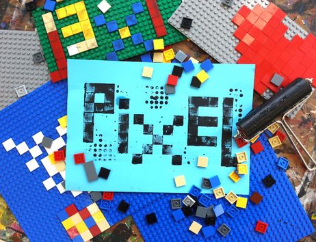 Bunte Steckbausteinplatten und lose Steine umgeben ein blaues Blatt mit dem gedruckten Wort „PIXEL“; rechts liegt eine Farbrolle.